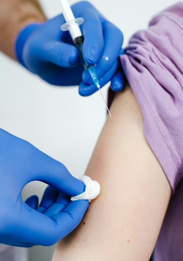 Healthcare worker wearing blue gloves administering a vaccine injection into a patient’s upper arm while holding a cotton pad.